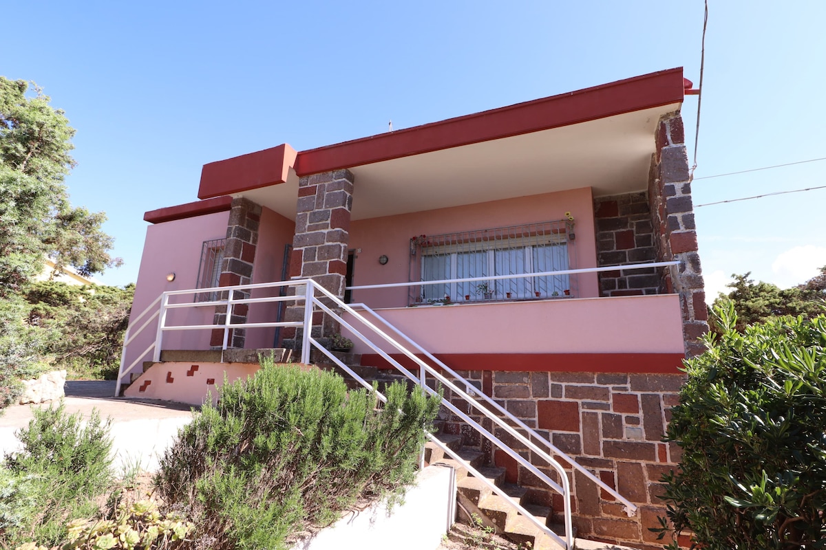 The exterior of the chalet features a welcoming stone façade with a pink finish and a red accent along the roof. A gently sloping path leads to the entrance, which is flanked by greenery and planters. A covered porch provides a space for relaxation.