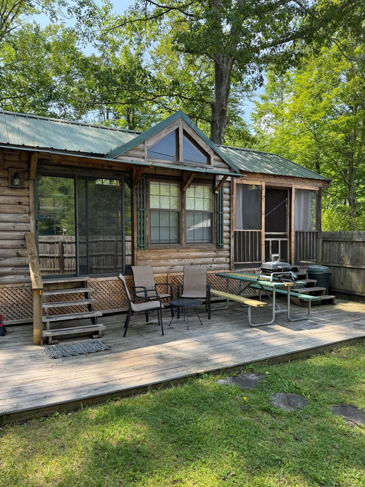 A cabin exterior is shown, featuring a green metal roof and wooden siding. A screened-in porch is visible, along with outdoor seating consisting of four chairs and a small table. Steps lead up to the entrance, surrounded by a well-maintained grassy area.