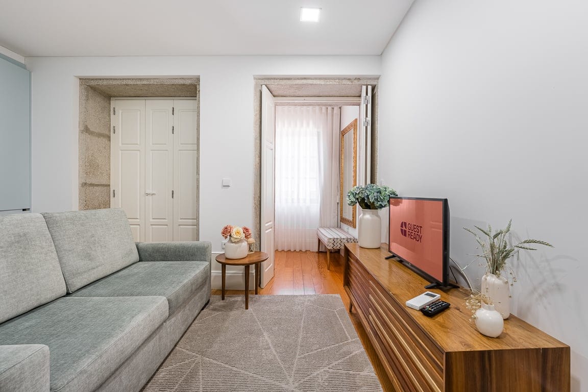 A modern living area is highlighted by a gray sofa facing a wooden console table with a television. Natural light enters through nearby windows, illuminating the space. Storage cabinets are visible in the background, adding functionality to the inviting layout.