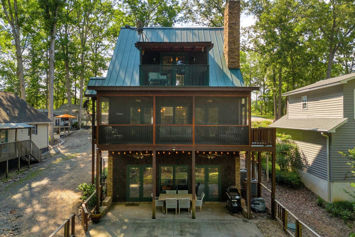 A multi-level house is surrounded by lush trees, featuring a charming combination of natural wood and stone accents. The lower patio area is equipped with outdoor seating, while the upper balcony showcases railings and string lights, creating a cozy spot for relaxation.