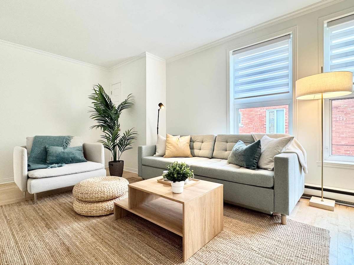 A bright living area is featured, showcasing a light-colored sofa adorned with throw pillows and a cozy armchair. A natural fiber rug complements a wooden coffee table, which is topped with a small potted plant. Sunlight enters through large windows, illuminating the space.