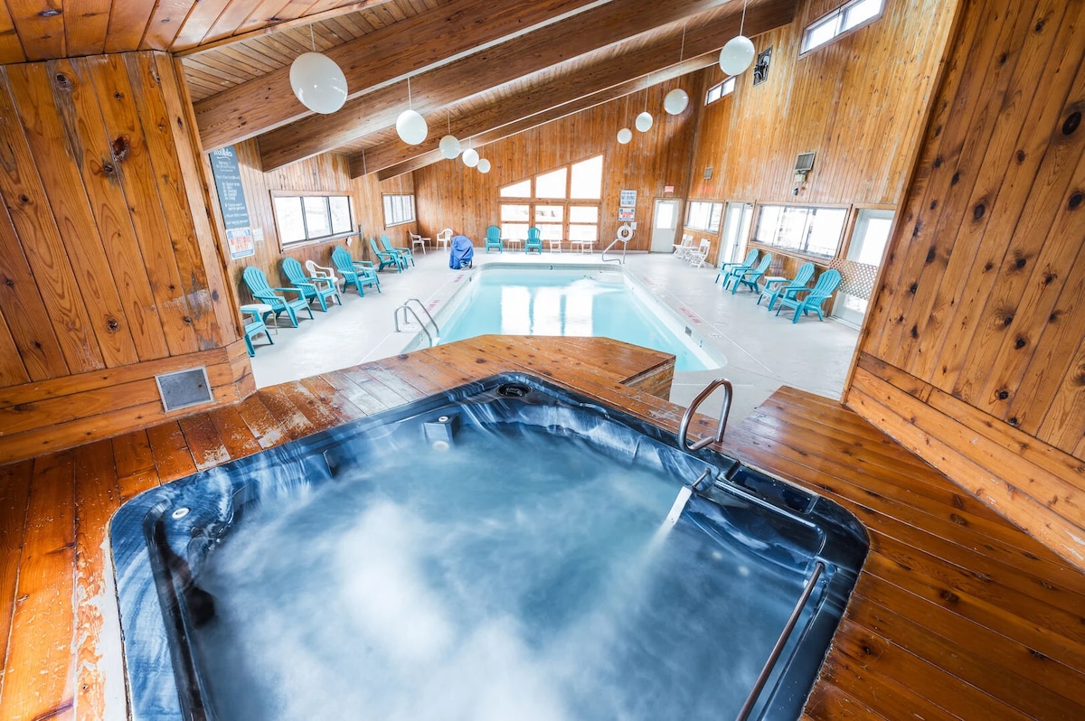 An indoor pool area is presented, featuring a hot tub in the foreground. Above, the wooden ceiling with large windows allows for ample natural light. Surrounding the pool are several lounge chairs, providing a comfortable space for relaxation.