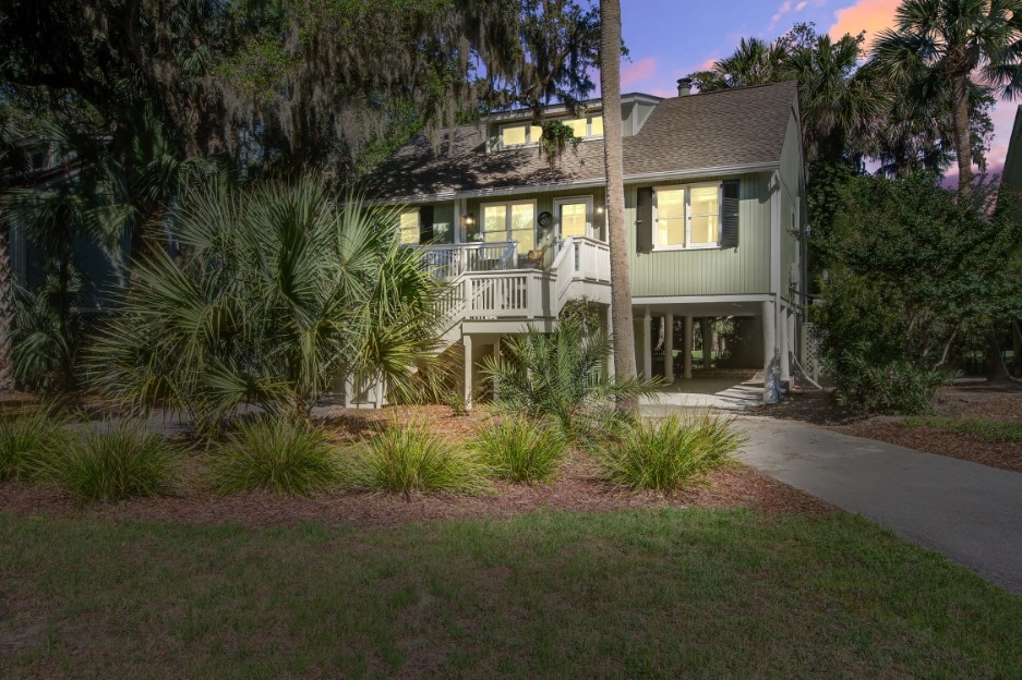 A charming beach cottage is set amidst lush landscaping, with palm trees and vibrant foliage surrounding the entrance. The home features a raised structure with a spacious porch highlighted by yellow shutters, while soft evening light enhances the inviting exterior.