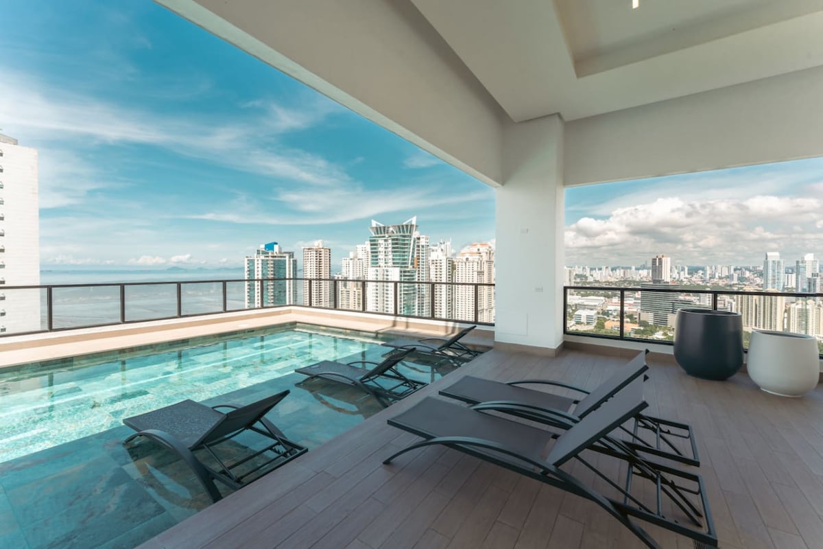 An outdoor terrace features a sleek infinity pool with stunning views of the Pacific Ocean and city skyline. Stylish lounge chairs are arranged around the pool, inviting relaxation in the expansive space under a bright blue sky.