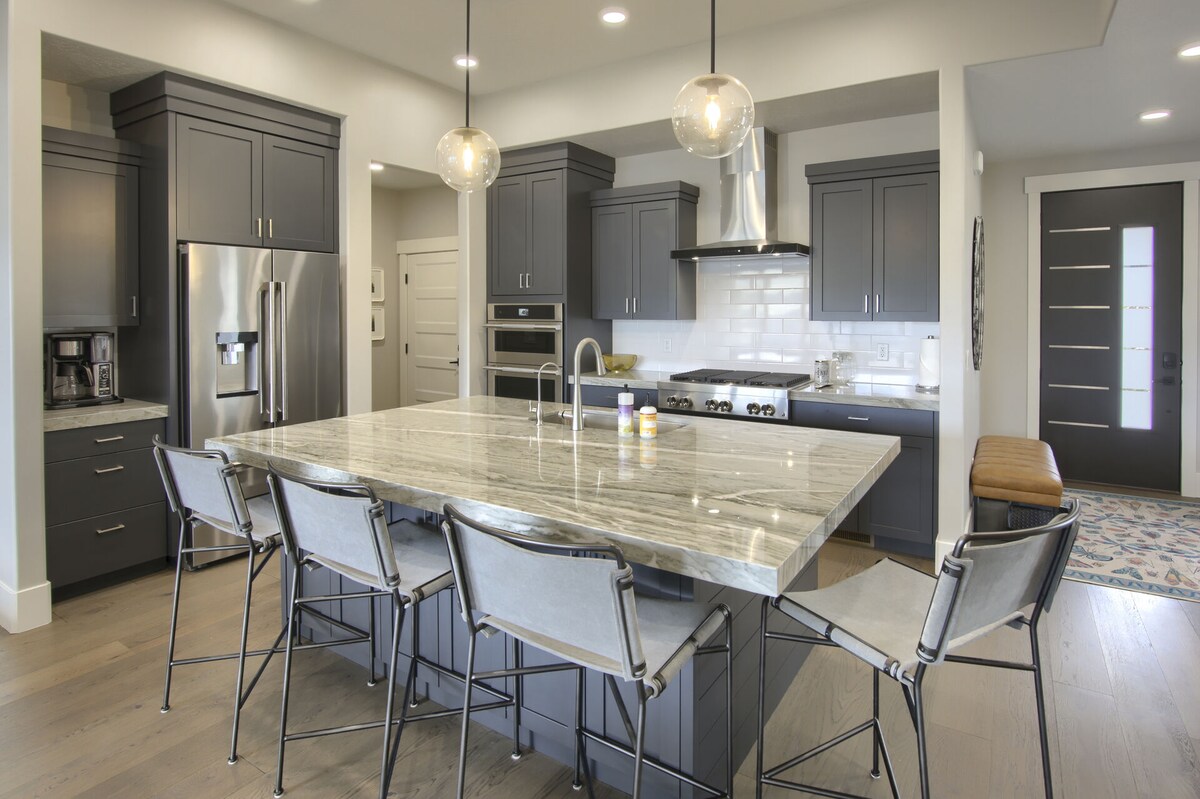 A modern kitchen features dark cabinetry and a spacious island with a marble countertop, complemented by seven metal barstools. Stainless steel appliances, including a refrigerator and oven, are visible, and the backsplash is white subway tile. Natural light enters through a nearby entrance door.