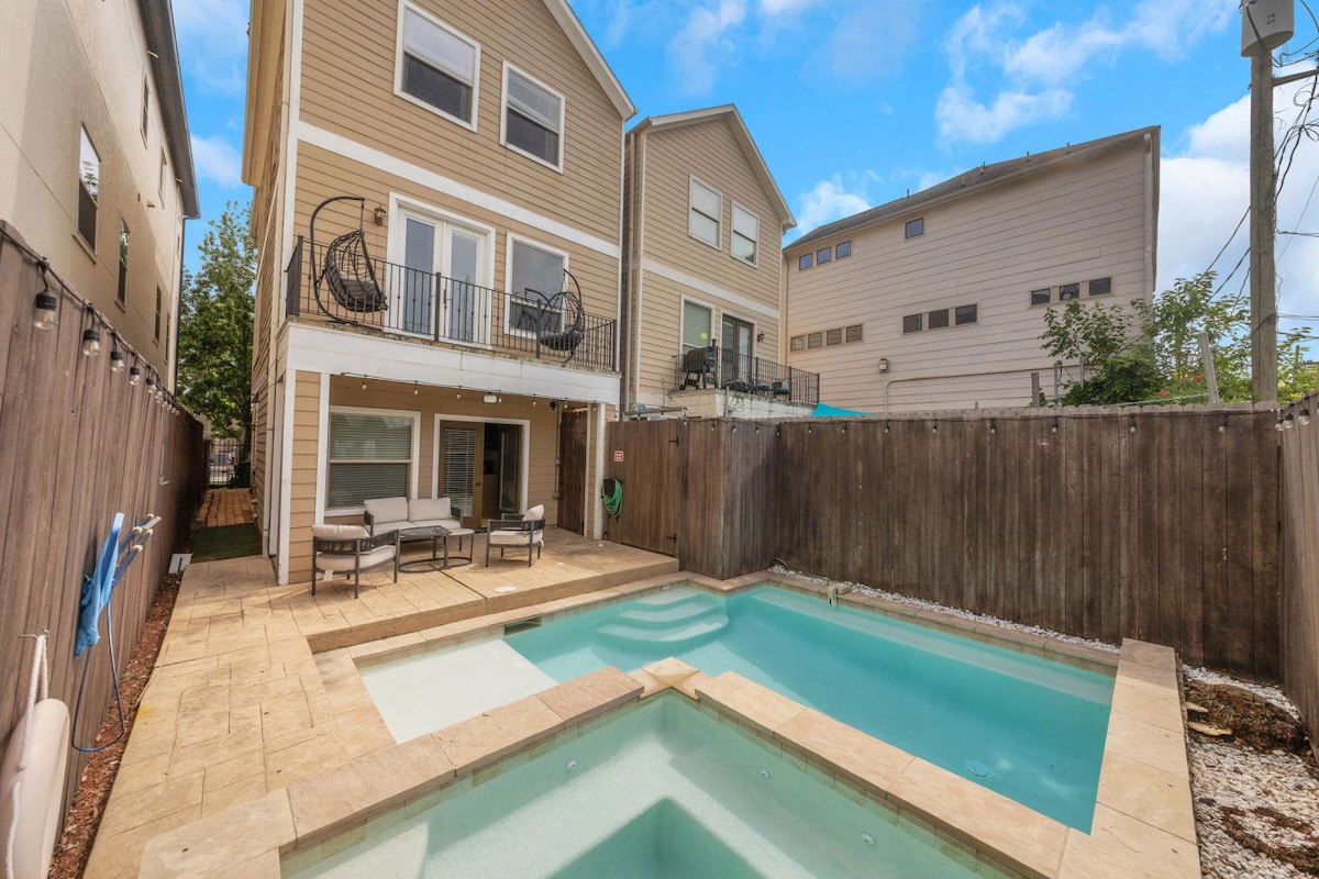 The outdoor area features a rectangular pool with a jacuzzi to the side, surrounded by a textured stone patio. The two-story home has multiple outdoor seating arrangements visible. A wooden fence ensures privacy, complemented by greenery in the background.