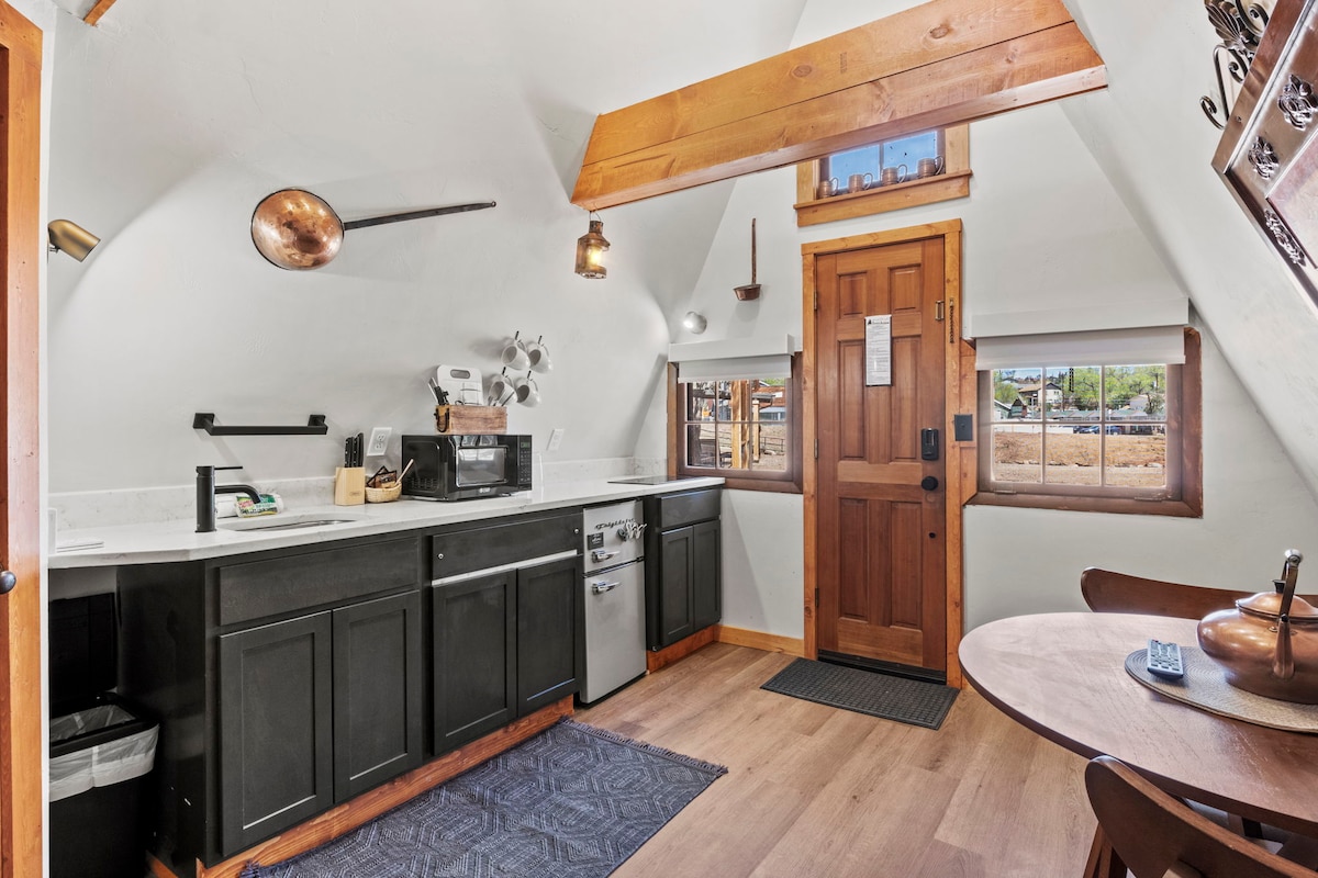 A compact kitchenette showcases modern appliances, including a microwave and refrigerator, under sloped ceiling beams. Dark cabinetry contrasts the light countertops, and a small dining table is positioned nearby. Natural light filters through the window, highlighting the inviting space.
