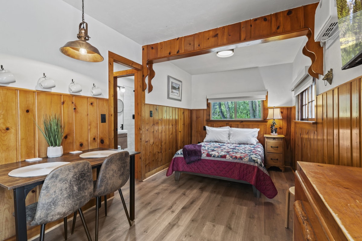 A cozy cabin interior features wood-paneled walls and warm lighting. A neatly made bed with a patterned quilt is positioned beside a window, offering natural light. A rustic dining table for two sits adjacent, with soft grey chairs complementing the space.
