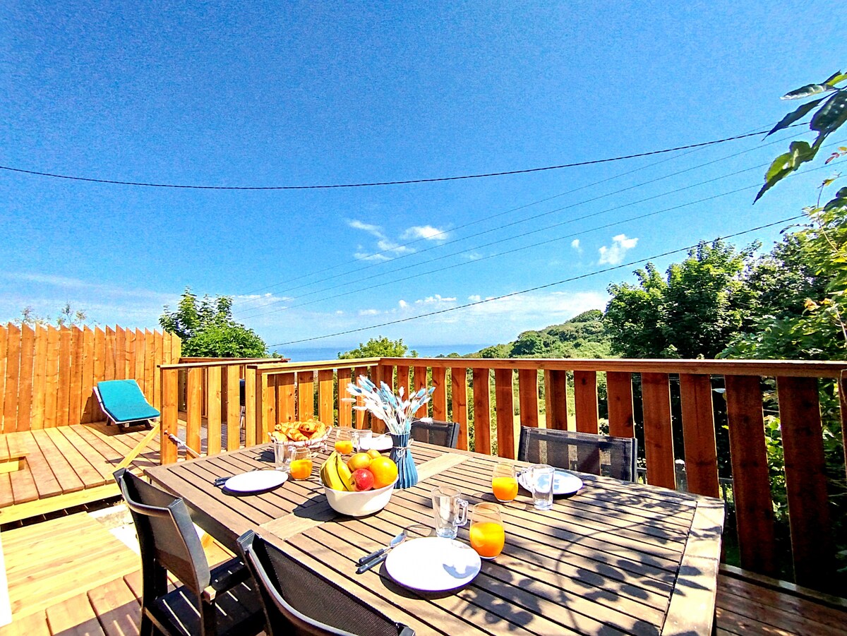An outdoor dining area is set on a wooden terrace, featuring a large table adorned with plates and glasses. A bowl of fresh fruit is present, along with orange drinks. Clear blue skies and greenery are visible in the background, enhancing the relaxed atmosphere.