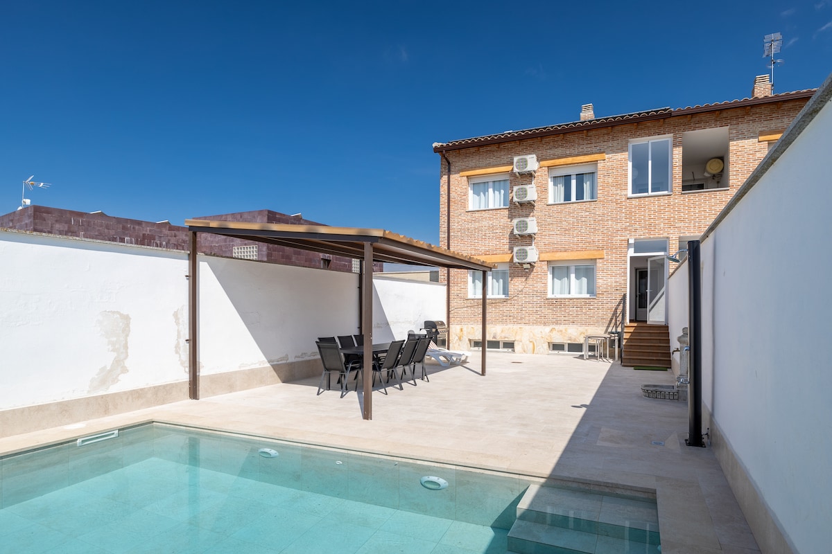An outdoor area is showcased with a private pool surrounded by a tiled terrace. A shaded seating area with multiple lounge chairs provides relaxation options. The brick structure of the house stands behind, featuring large windows that allow natural light to enter the interior.