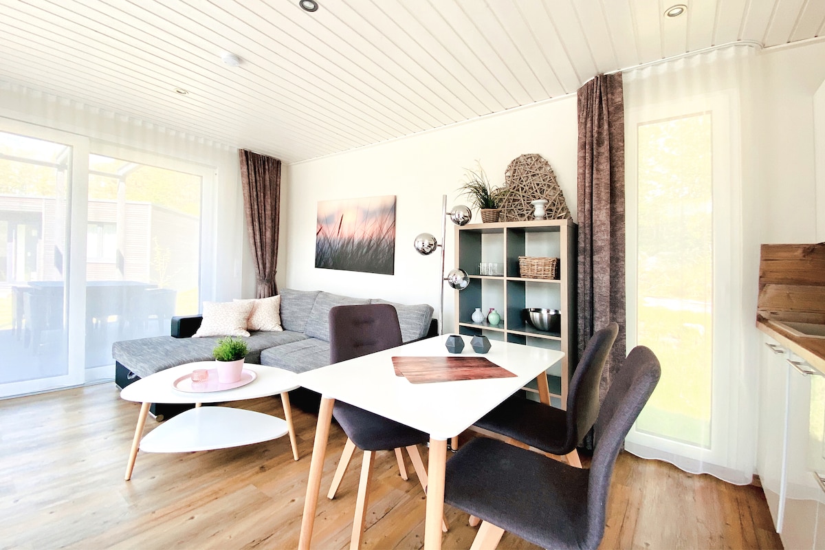 The living area features a modern design with a spacious gray sofa facing a dining table. A light wood floor complements the room. A decorative shelving unit holds various items, and large windows allow natural light to fill the space. Artwork is displayed on the wall.
