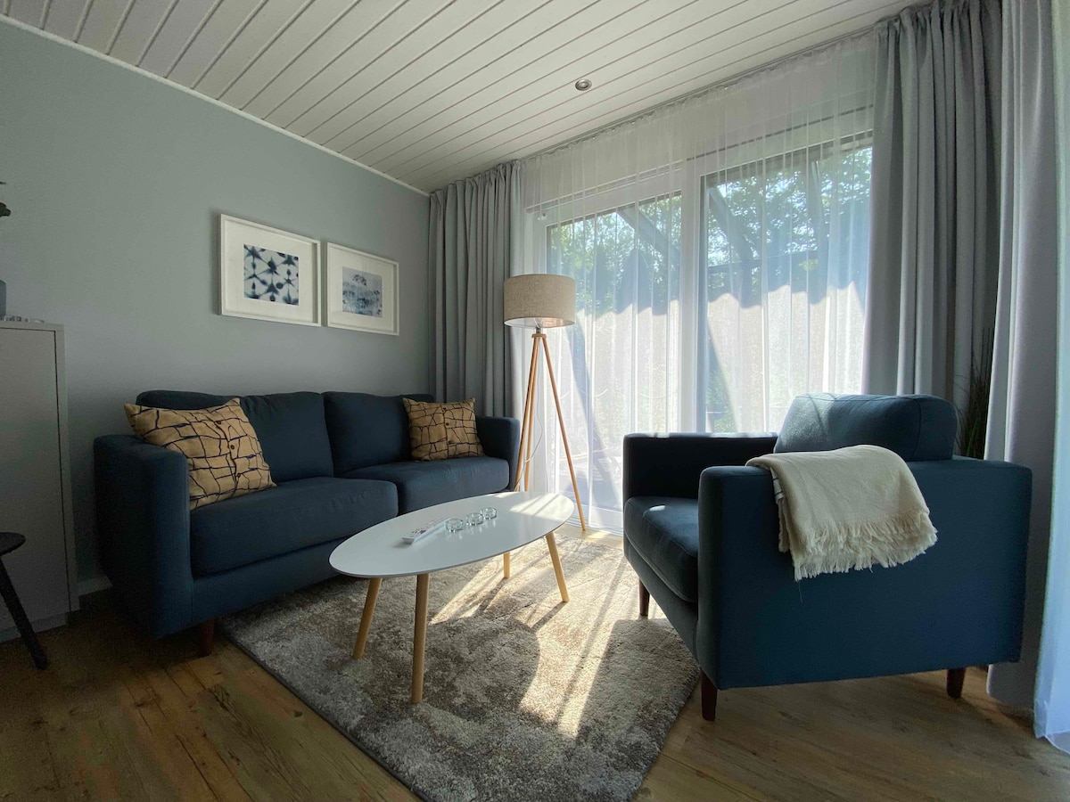 A modern living area is presented with a dark sofa and two matching armchairs. A light coffee table is centrally located on a soft area rug. Natural light filters through sheer curtains, illuminating the space and highlighting the light tones of the wooden flooring.