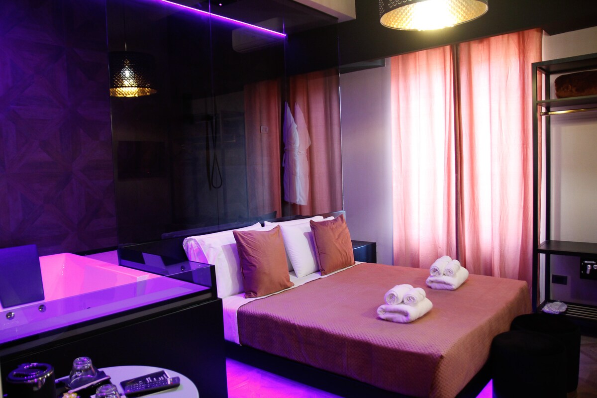 A king room features a large bed with plush pillows and neatly arranged towels. Soft lighting highlights the modern decor, while floor-to-ceiling windows with sheer curtains add a sense of openness. A private bathtub and clothing storage area are visible in the well-appointed space.