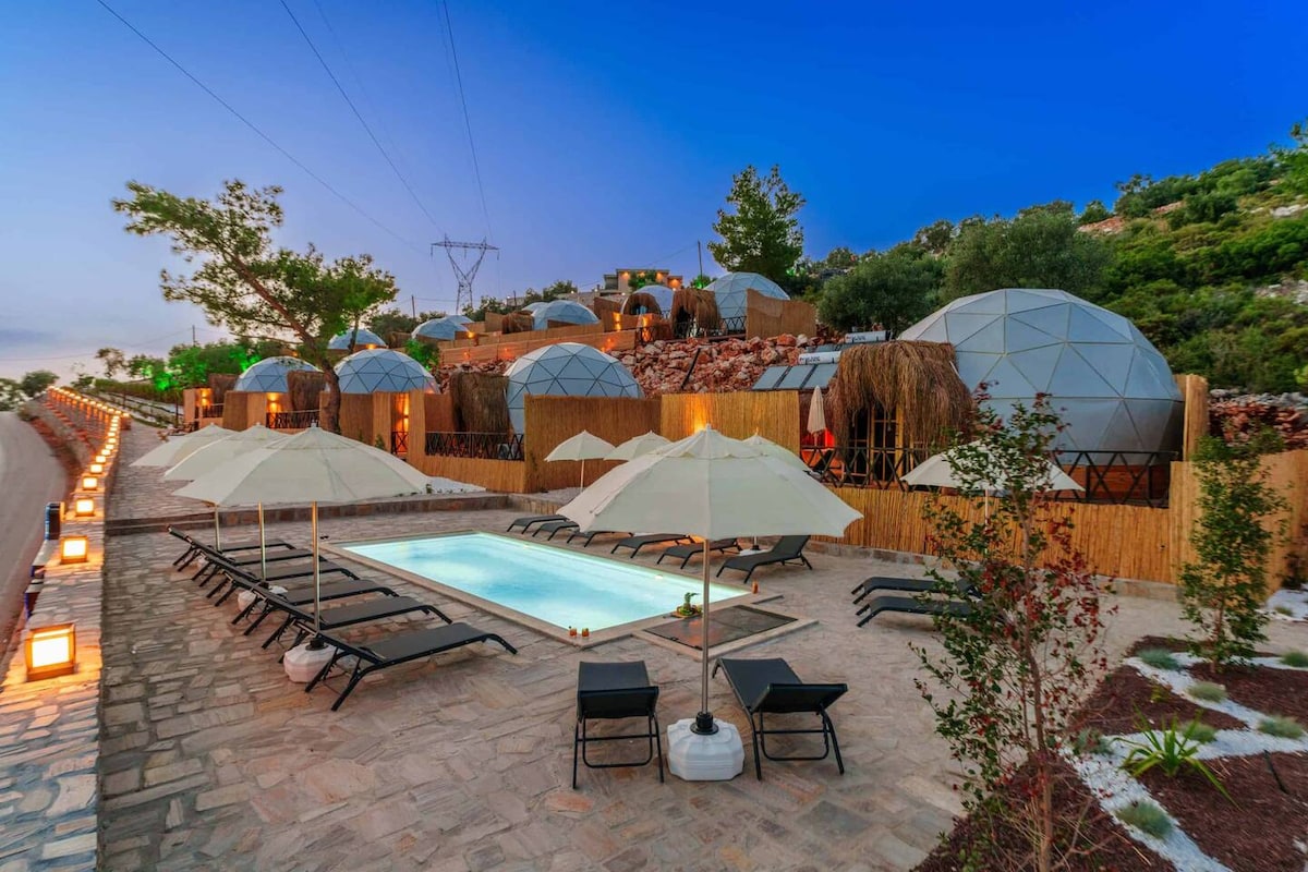 The outdoor area showcases a private pool surrounded by lounge chairs and umbrellas. Lush greenery and carefully arranged stones create a natural setting. Multiple dome structures are visible in the background, illuminated softly as evening approaches, highlighting a serene and exclusive atmosphere.