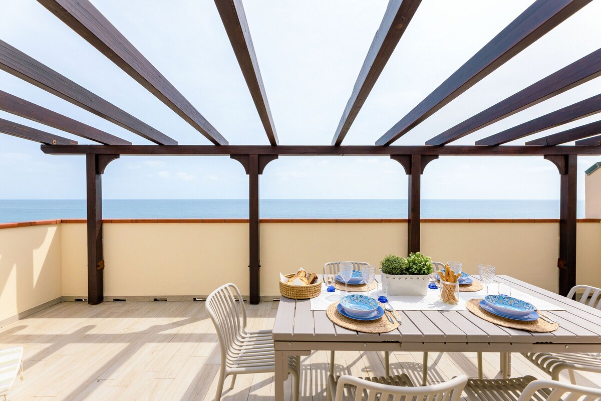 An expansive private terrace offers a dining table set for outdoor meals. The area is shaded by wooden beams, with a direct view of the Adriatic Sea visible in the background. Light-colored flooring complements the coastal atmosphere while inviting relaxation and enjoyment.