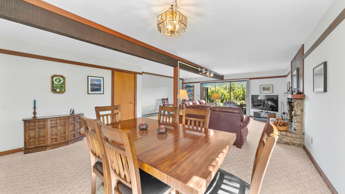 An inviting dining area features a large wooden table surrounded by six chairs, with a subtle warm wood finish. Natural light streams through sliding glass doors, illuminating the open space that connects the dining area to a cozy living room with a stone fireplace.