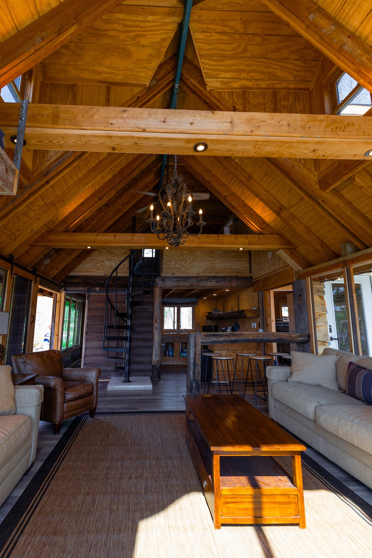 An inviting open-layout cabin is showcased with high, wooden ceilings. Light filters through large windows onto a spacious living area featuring a coffee table and comfortable sofas. A spiral staircase is visible, leading to an upper level, while a chandelier enhances the rustic atmosphere.