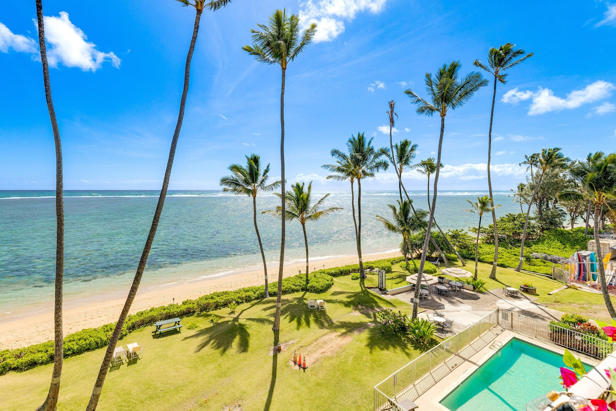 A panoramic view of the ocean and sandy beach is framed by swaying palm trees. A grassy area is visible, featuring picnic tables and a swimming pool. The clear blue sky enhances the tranquil setting, perfect for enjoying the natural beauty of the coastline.