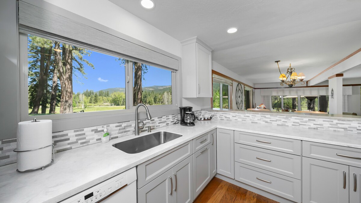 The kitchen features white cabinetry, a marble countertop, and a spacious sink. Large windows allow natural light to brighten the space, with views of surrounding trees and greenery visible. Modern appliances, including a coffee maker and dishwasher, are arranged neatly, enhancing functionality.