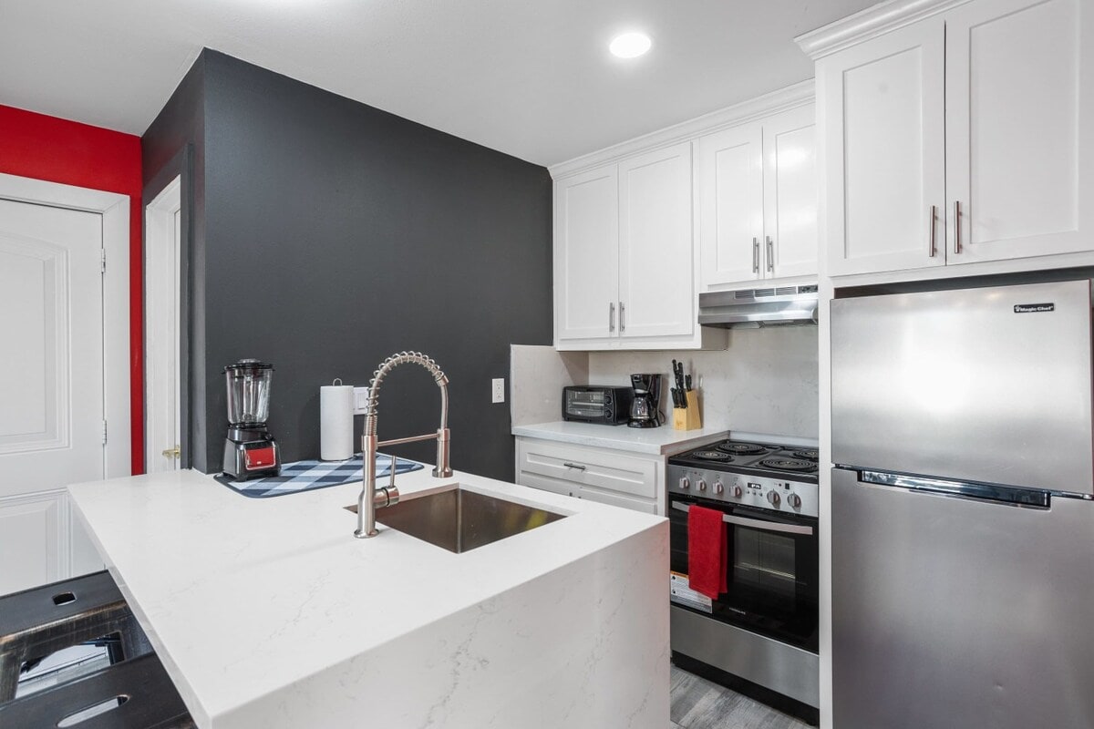 A modern kitchen is presented, featuring white cabinetry and a contrasting dark wall. Stainless steel appliances include a refrigerator and gas stove. A central island with a sink serves as a functional workspace, complemented by a blender and dish towels on the countertop.