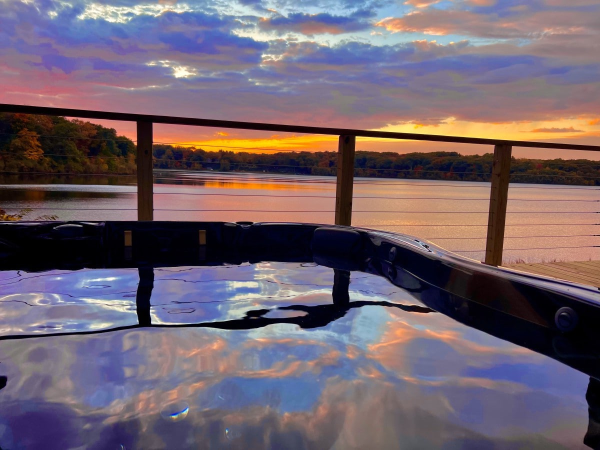 A hot tub sits on a deck overlooking Stone Lake, reflecting the vibrant colors of the sunset in its still water. The sky is filled with hues of orange, pink, and purple, providing a serene backdrop to the tranquil lake and surrounding trees.