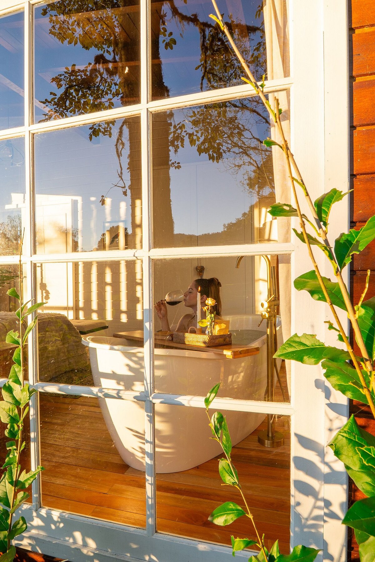 A spacious bathtub is visible through a large window framed by wooden panes, reflecting natural light and greenery. A small tray rests atop the tub, showcasing a candle and decorative pieces. The warm wooden floor adds a touch of coziness to the setting.