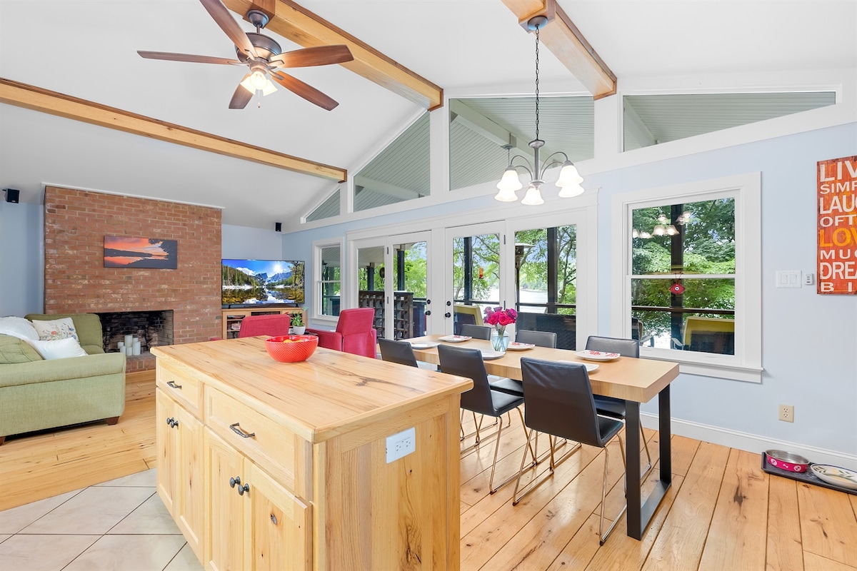 An inviting interior space with vaulted ceilings and warm wood accents is showcased. A central dining area features a wooden table surrounded by black chairs, while a kitchen island offers additional counter space. Large windows frame a view of the lake, creating a bright atmosphere.