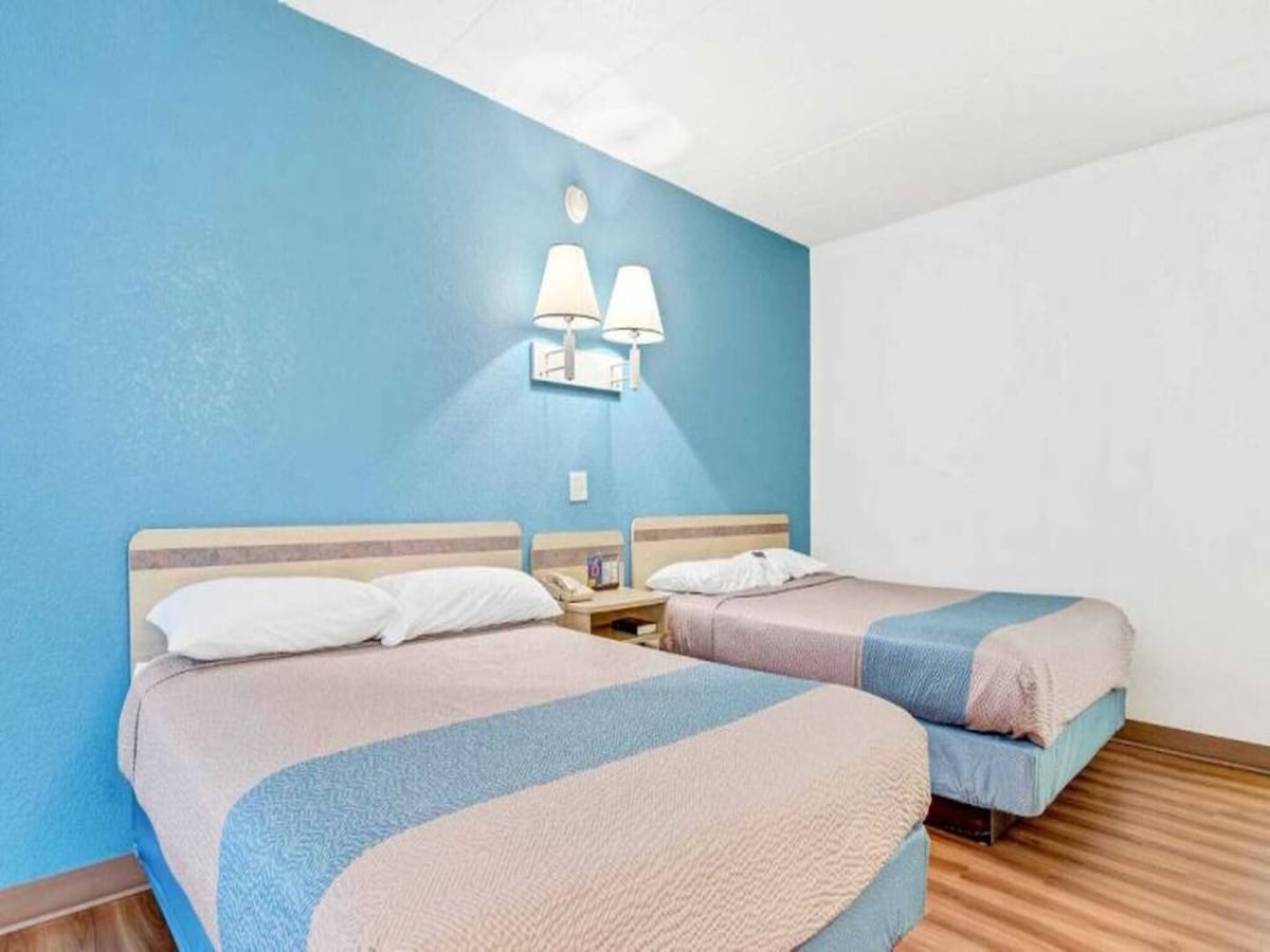 A bright room features two queen beds with light bedding, positioned against a blue accent wall. Each bed is accompanied by a nightstand with wall-mounted lamps, providing soft illumination. The wood-style flooring adds warmth to the space.
