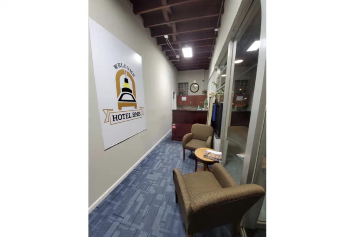 A hallway features a welcome sign for the hotel along with a reception area visible in the background. Two brown chairs are positioned near a small table, creating a comfortable seating area. Great natural light filters in through large windows, enhancing the space.