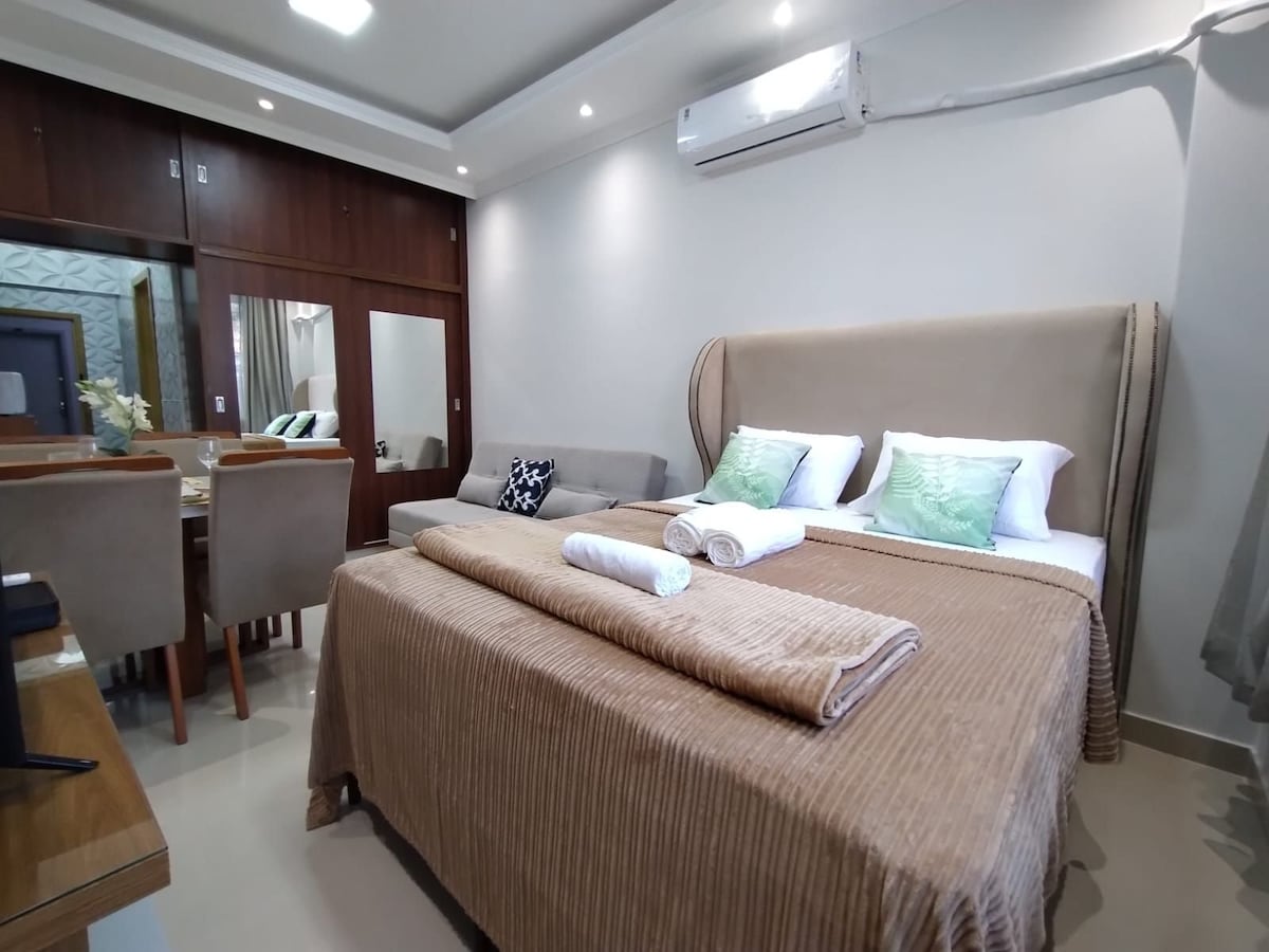 A comfortable sleeping area features a spacious bed with soft bedding and decorative pillows. Light walls create a bright atmosphere, while a dining table with chairs is visible in the background. Air conditioning is mounted above, adding to the convenience of the space.