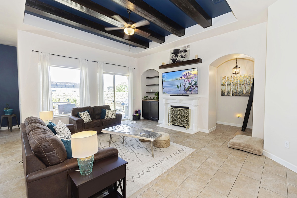 An open-concept living area features a spacious seating arrangement with dark sofas and a decorative coffee table. Large windows allow natural light to fill the room, accented by vaulted ceilings and ceiling beams. A television is mounted above a modern fireplace, enhancing the inviting atmosphere.