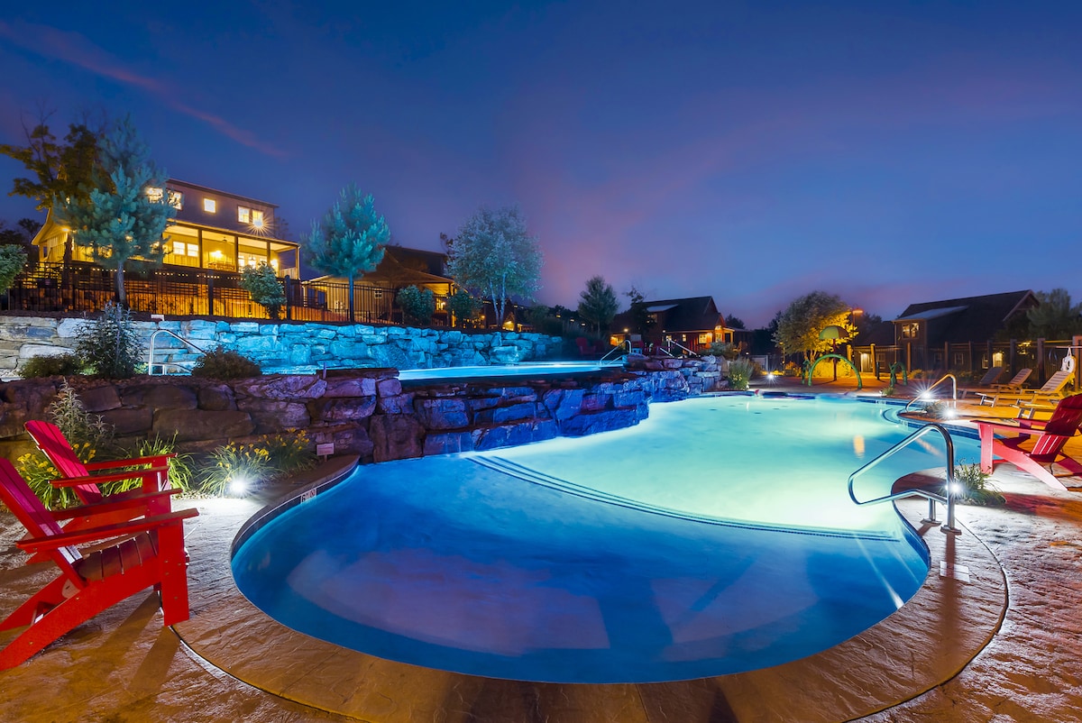 The image showcases a serene pool area illuminated under the night sky. The pool features gentle curves, with softly lit water and surrounding rock formations. Red adirondack chairs are positioned nearby, inviting relaxation and enjoyment of the tranquil atmosphere.