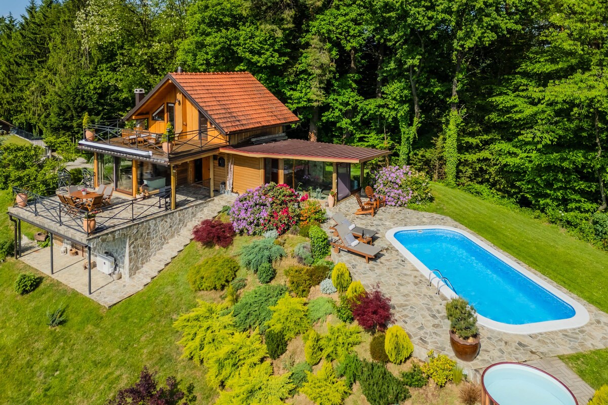 A modern holiday home is nestled amid lush greenery, featuring a private outdoor pool surrounded by landscaped gardens. A spacious deck with dining and lounging areas offers scenic views, while vibrant flowers add color to the serene environment.