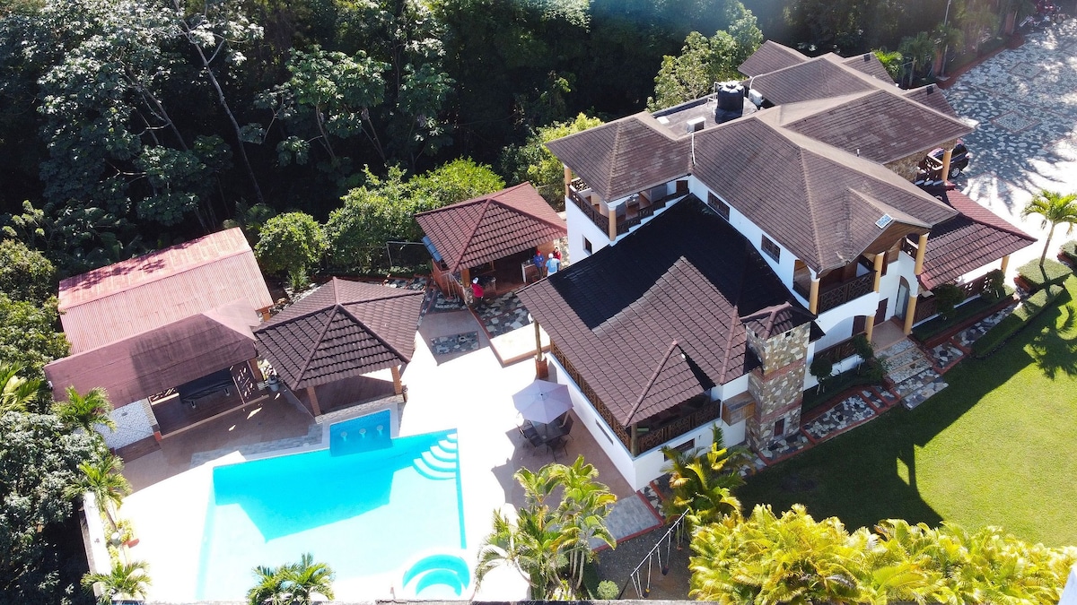 An aerial view of the spacious property showcases a heated pool surrounded by tropical greenery. Multiple patios and outdoor seating areas provide ample space for relaxation. The multi-level structure features a diverse roofline and is complemented by nearby outdoor kitchens and barbecue facilities.