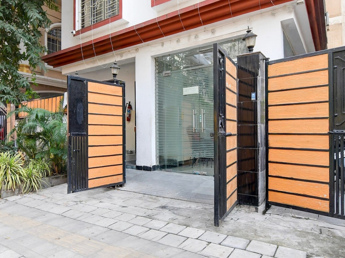 The entrance features a pair of wooden gates, bordered by low greenery and a stone pathway. A large window is visible behind the gates, allowing natural light into the interior. The modern design showcases a blend of orange and black elements.