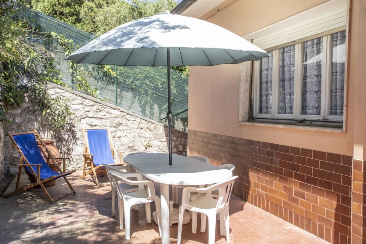 An outdoor area is featured with a round white table surrounded by four chairs, providing ample space for dining. A large umbrella offers shade, and two blue lounge chairs are positioned nearby. The wall is adorned with textured brown tiles, and greenery is visible in the background.