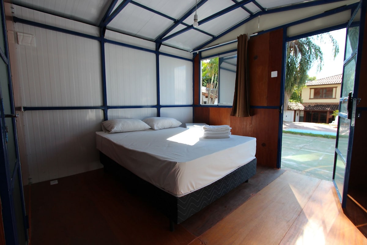 A bright and airy sleeping area is furnished with a spacious bed topped with rolled towels. Large windows allow natural light to fill the room, while minimalistic decor creates a simple and functional ambiance. Window treatments provide an option for privacy.