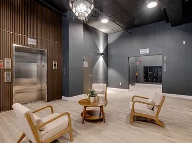 The modern lobby features a sleek design with a combination of wooden accents and dark walls. An elevator is positioned prominently, while a small circular table is surrounded by two light-colored chairs, creating a welcoming seating area.