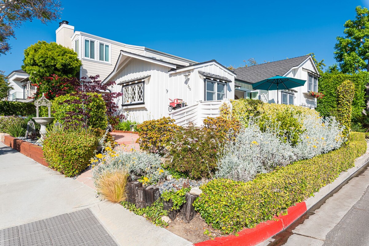 A charming cottage is showcased at the corner lot, surrounded by an array of colorful shrubs and flowering plants. A landscaped path leads to the front steps, with a solid seating area across the property. The clear blue sky enhances the inviting atmosphere.