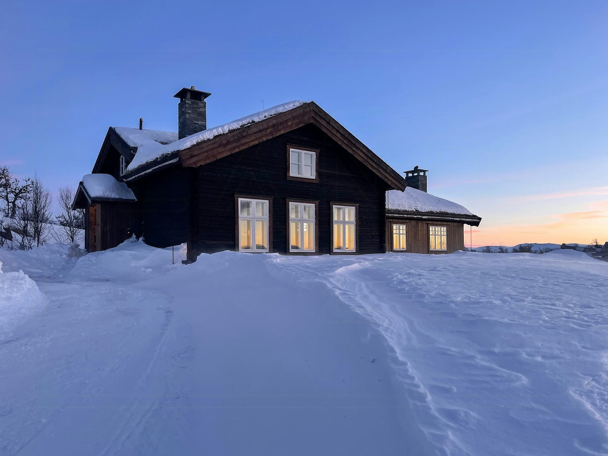 A charming cabin is viewed against a backdrop of a wintry landscape, with snow covering the ground. Soft light glows from the windows, creating a cozy atmosphere as the sun sets in the distance, merging warm hues with the cool colors of the snow.