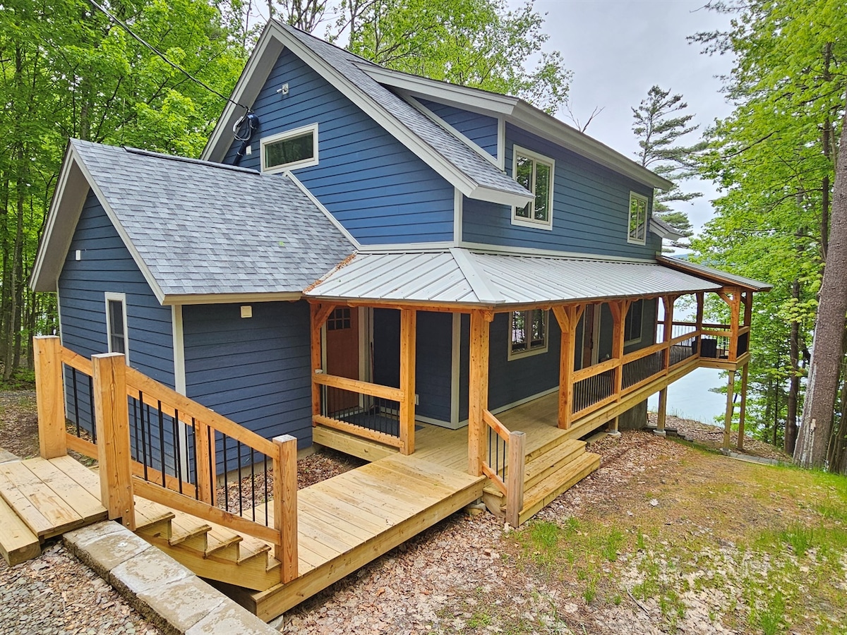 The exterior of a newly constructed post and beam home is displayed, featuring a combination of dark blue siding and a metal roof. A wooden deck extends around the home, providing access to outdoor spaces surrounded by lush greenery.