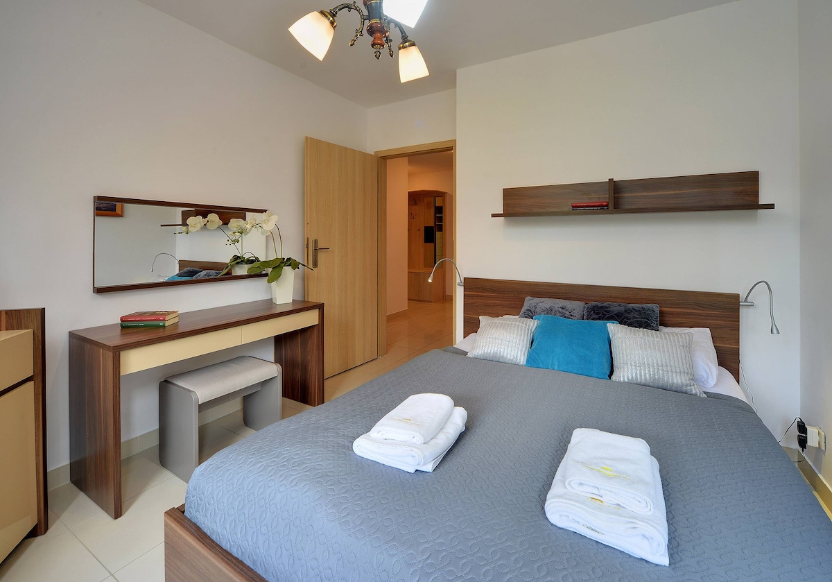 Një dhomë gjumi me një krevat dopio të madh, dy tavolina pranë krevatit dhe një pamje të një kuzhine të vogël. Shtrojat e buta dhe një sistem minimalist i japin dhomës një atmosferë të qetë dhe të thjeshtë.