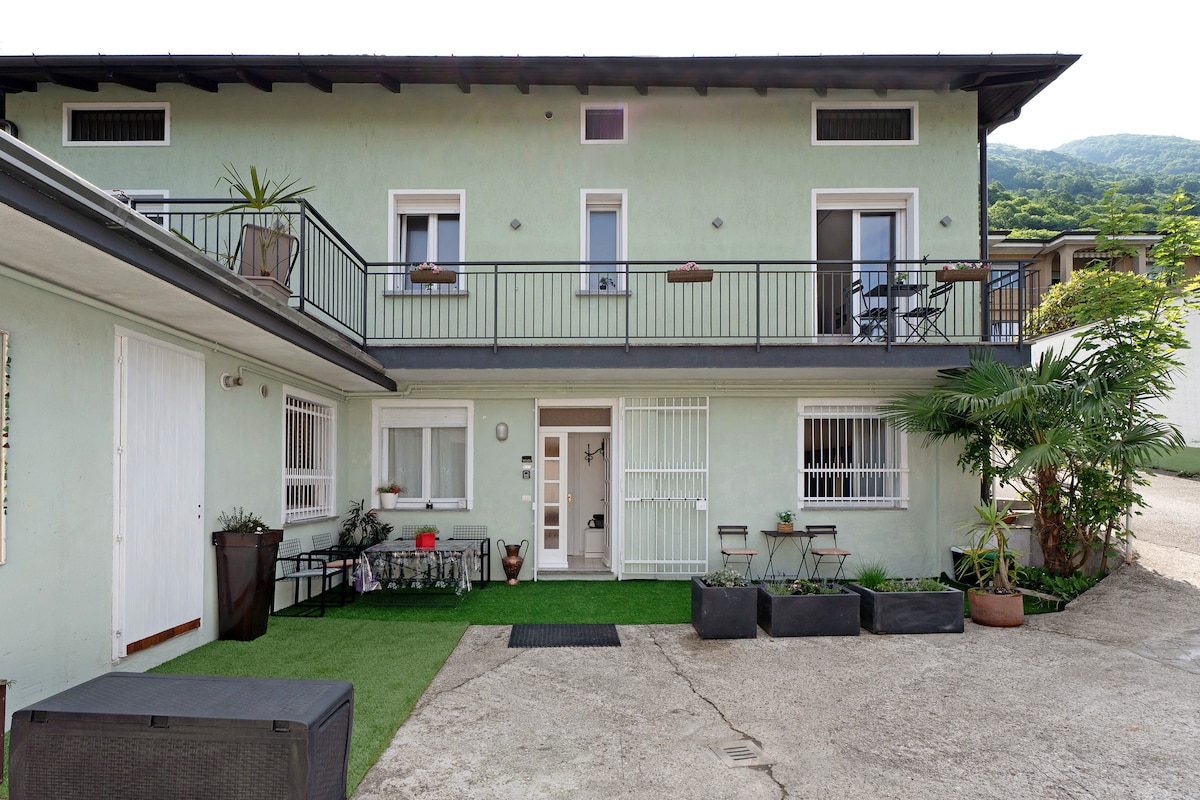 The exterior of Casa Lago Leo displays a step-free, welcoming entrance surrounded by greenery. A small patio area features potted plants and two seating arrangements, while the upper balcony offers additional outdoor space. The structure is complemented by the serene mountain backdrop.