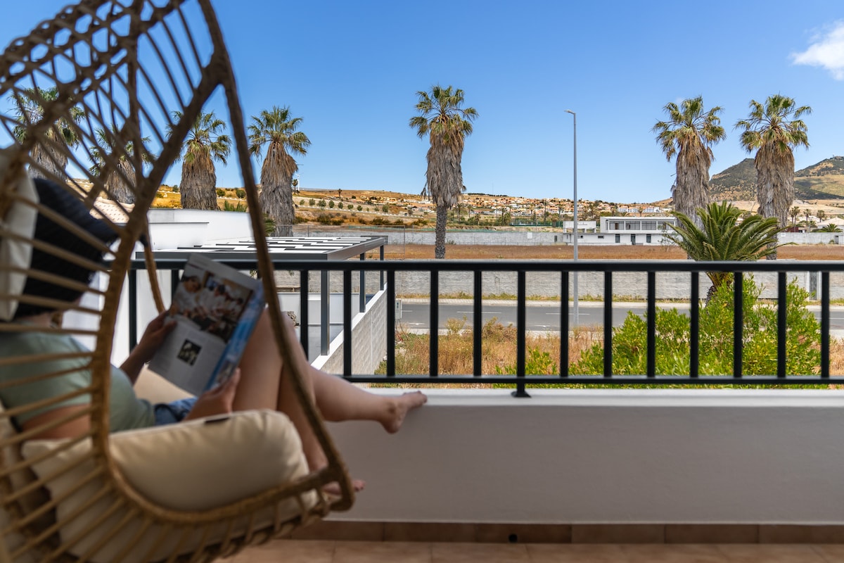 A cozy balcony offers a view of palm trees swaying gently, with a spacious area visible beyond. A person sits comfortably in a hanging chair, engaged in reading. The bright blue sky enhances the relaxing atmosphere.