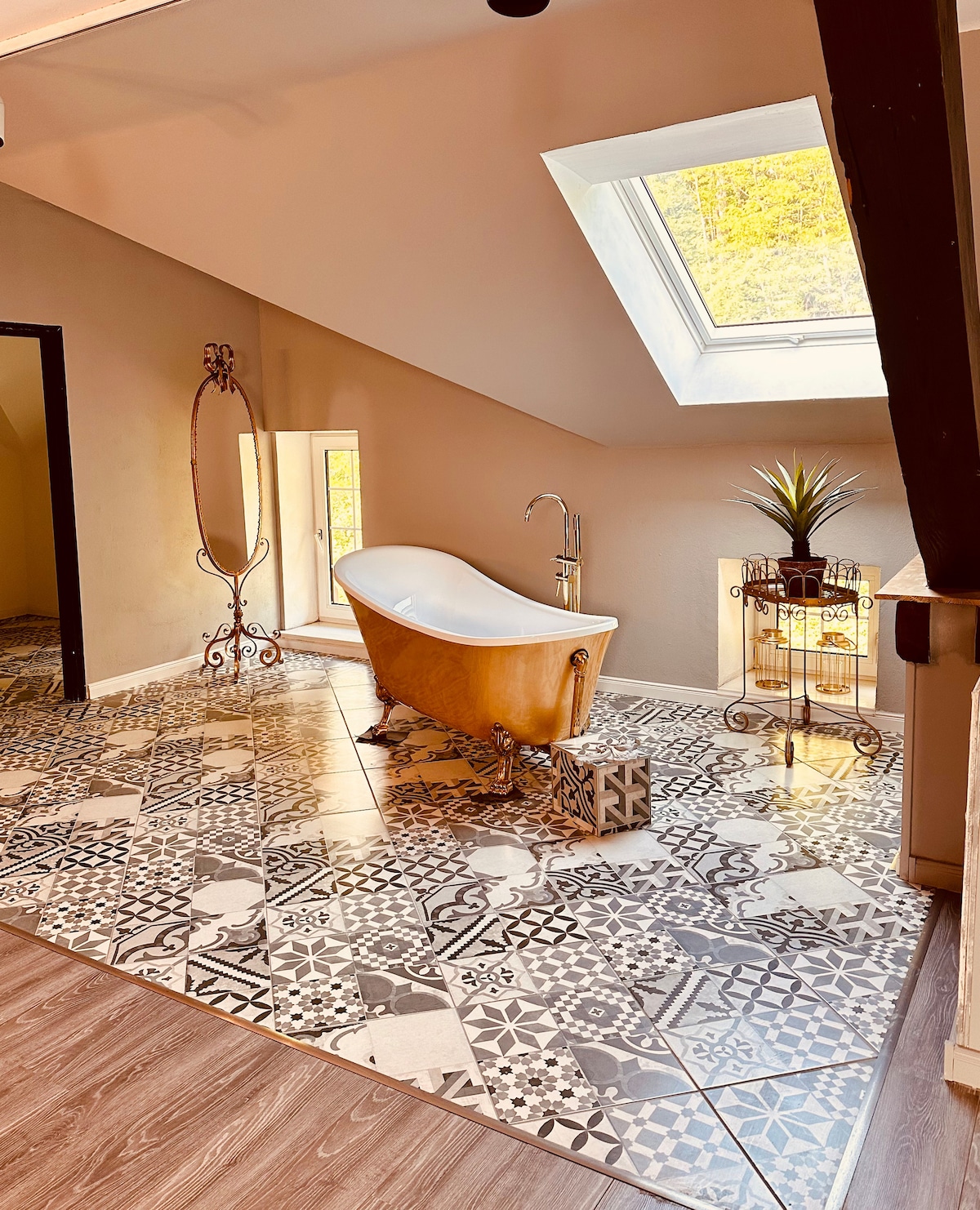 A spacious bathroom is highlighted by a freestanding bathtub positioned near a skylight. The floor features intricate patterned tiles, and a decorative plant adds a touch of greenery. A vintage-style mirror stands in the corner, complementing the room's design.