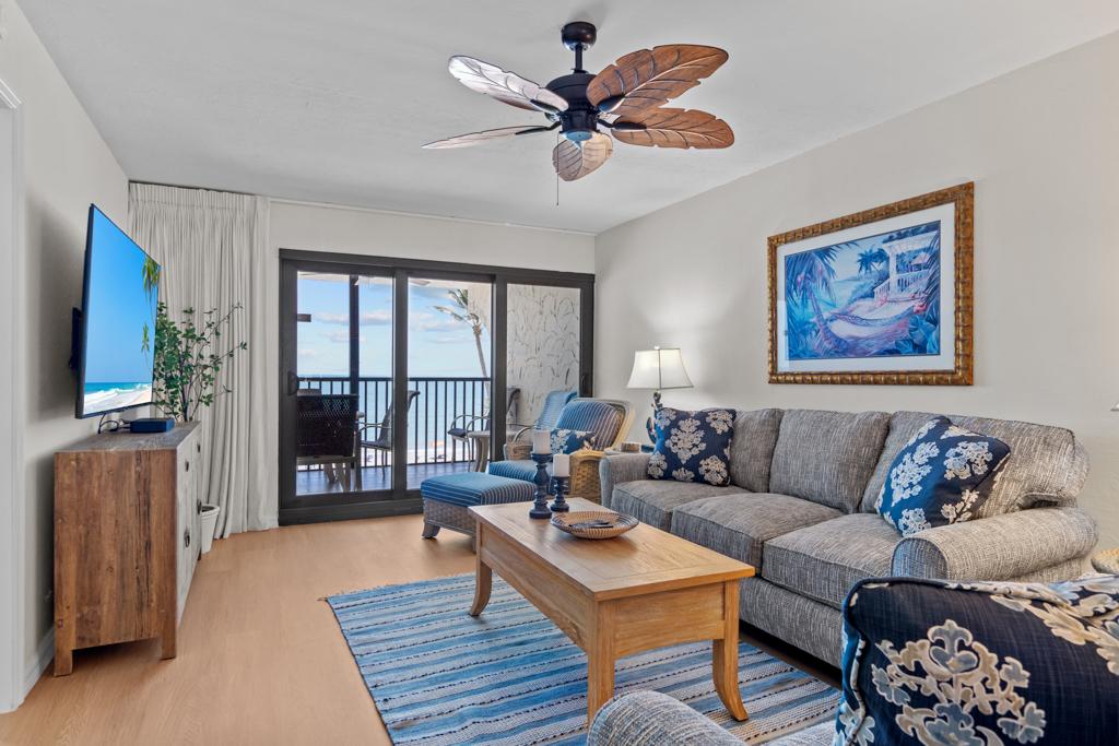 A comfortable living area features a large sectional sofa adorned with coastal-themed cushions. A wooden coffee table sits at the center, with a TV mounted on the wall opposite. Sliding glass doors open to reveal a balcony showcasing views of the Gulf.