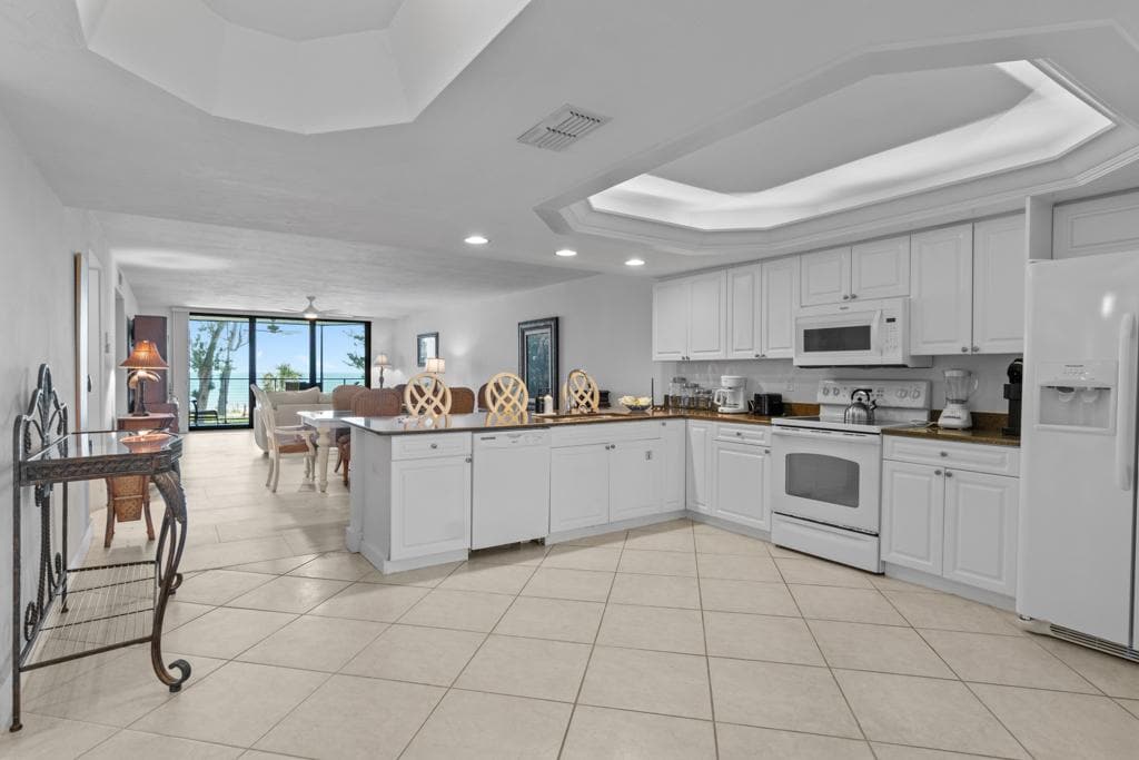 A spacious kitchen area features white cabinetry and a large island with three high-backed stools. Natural light flows through sliding glass doors, which lead to a living area with a view of the Gulf. Neutral tile flooring connects the kitchen and living spaces.