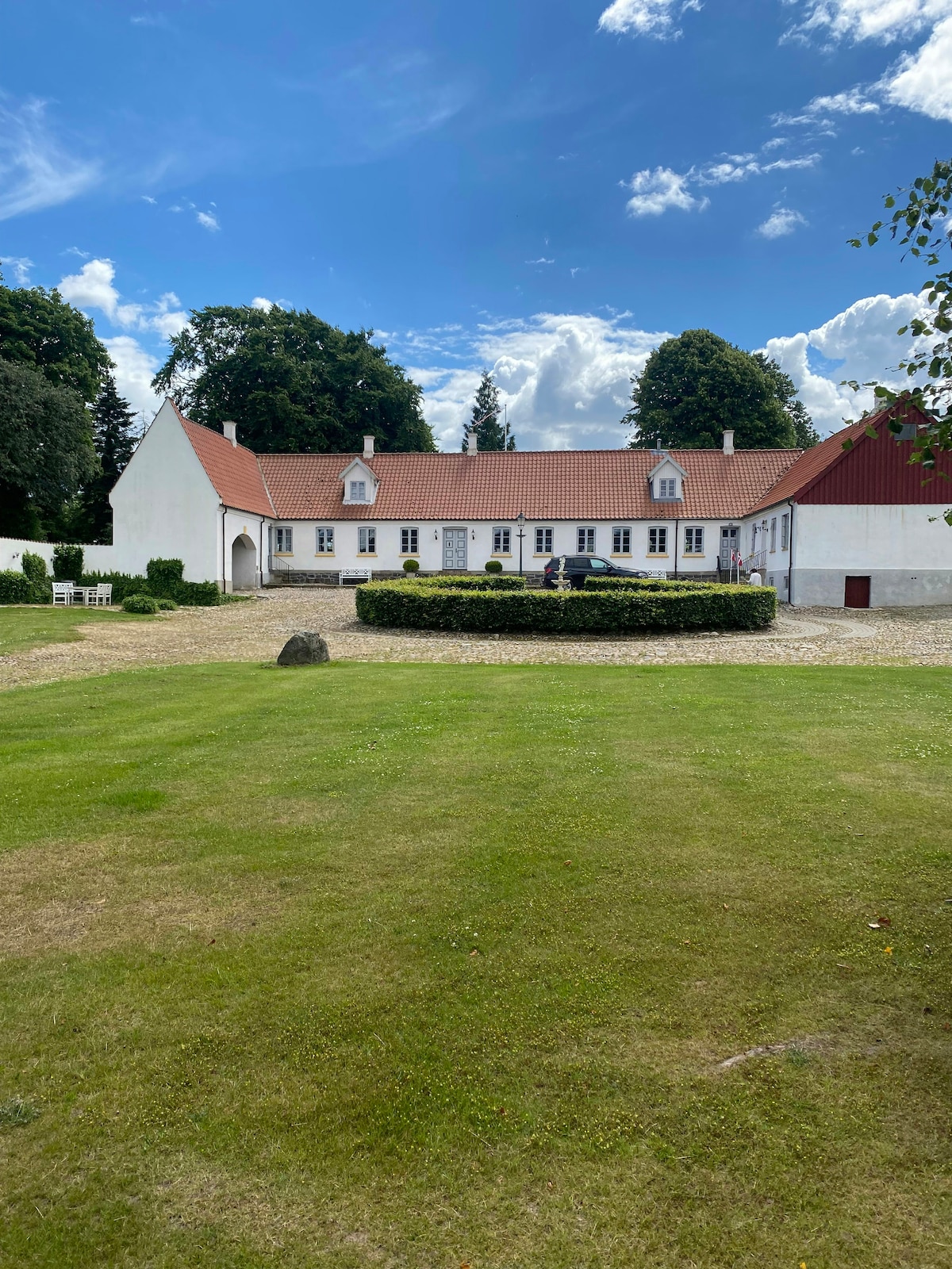 The farmhouse is displayed against a bright sky, showcasing its traditional architecture with white walls and a red roof. A neat lawn extends before the house, surrounded by well-maintained hedges, and a large stone is placed in the grassy area.