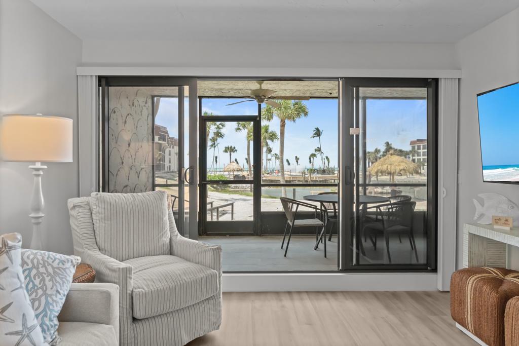 The living area displays comfortable seating with a textured armchair positioned near large sliding glass doors. These doors open to a patio with outdoor seating, providing views of palm trees and beach amenities.