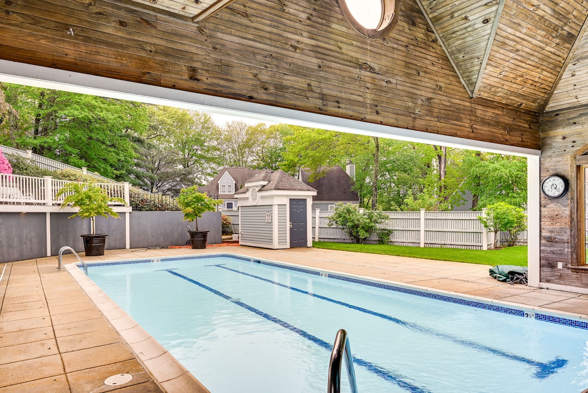 An indoor pool area features a clean, blue water pool beneath a wooden vaulted ceiling. Large windows allow natural light to flood the space, revealing greenery outside. The pool deck is bordered by potted plants, providing a touch of nature indoors.